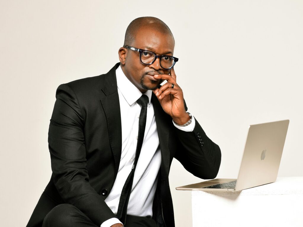 Confident businessman in a suit and glasses sits with laptop, hand on chin, showcasing professionalism.
